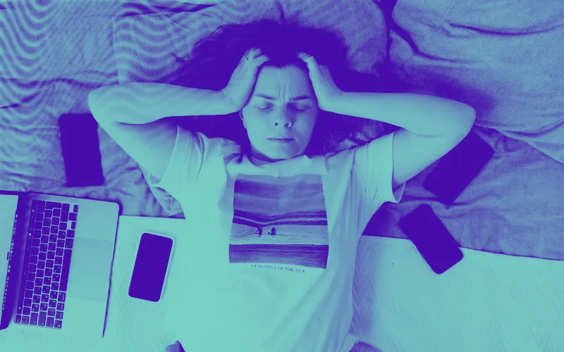 mulher deitada em cima da cama com computador e vários dispositivos móveis, com as mãos na cabeça em sinal de preocupação