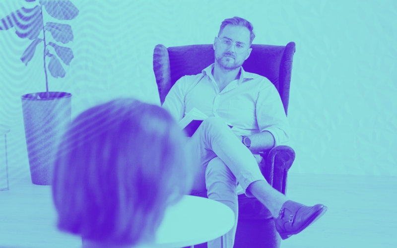 imagem de uma terapeuta com um bloco de apontamentos e uma pessoa que está à sua frente num ambiente clínico